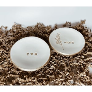 Pode incluir: Dois pratos de cerâmica branca para anéis com detalhes dourados. Um prato tem as iniciais "C & M" com um símbolo de coração entre as letras. O outro prato tem um desenho floral dourado e o nome "JANE" escrito em dourado.