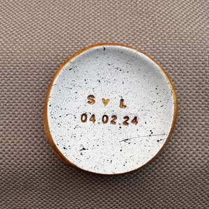 Pode incluir: Prato de cerâmica branca para anéis com borda dourada e manchas pretas. O prato tem as iniciais "S♥L" e a data "04.02.24" escritas em ouro.