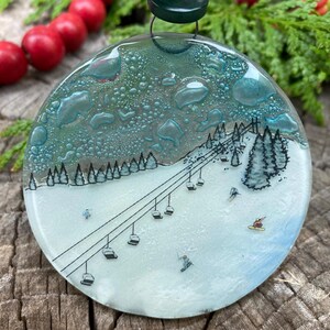 May include: Round glass ornament with a winter ski slope scene. The ornament features a ski lift, skiers, and snow-covered trees. The top has a textured, teal-colored design. Red berries and evergreen branches are in the background.