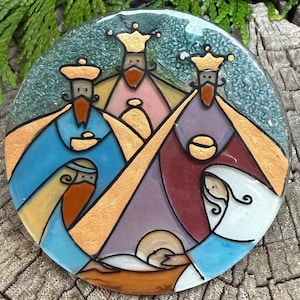 May include: A round, stained-glass ornament depicting the three wise men, Mary, and baby Jesus. The figures are in blue, gold, purple, and white. The ornament is on a weathered wooden surface.