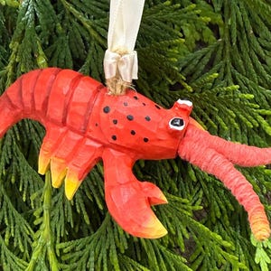 May include: A hand-carved, red lobster ornament with black spots and yellow accents. The lobster has a cream-colored ribbon for hanging. The ornament is set against a backdrop of green foliage, suggesting a nautical or coastal theme.