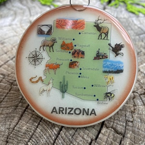 May include: A round, white, glass ornament with a map of Arizona on it. The map is green and brown with blue dots for cities. The cities are labeled with their names. The ornament has a red, white, and blue ribbon attached to it. The ornament is on a wooden surface.