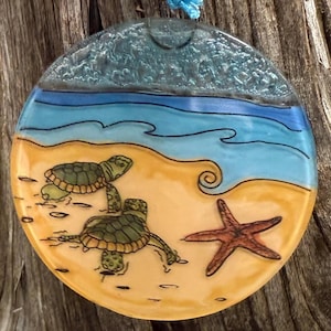 May include: Round, hand-painted glass ornament featuring a beach scene. The design includes two green sea turtles on a sandy shore, a brown starfish, and blue ocean waves. The top has a textured, light blue section representing the sky.