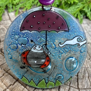 May include: A glass ornament with a blue background and a ladybug holding an umbrella. The ladybug is red with black stripes and white spots. There are bubbles and a cloud in the background.