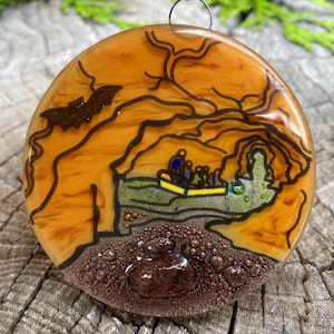 May include: A round glass ornament with a brown, orange, and yellow background. The ornament features a black silhouette of a bat flying over a cave with people inside. The cave is surrounded by green and brown foliage. The ornament has a brown, textured bottom with air bubbles.