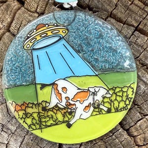 May include: A round, colorful pendant featuring a cow being abducted by a UFO. The design includes a yellow and orange UFO, a blue tractor beam, a white and orange cow, and a green and yellow landscape. The pendant has a blue string.