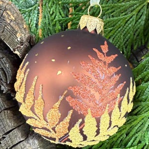 May include: A brown Christmas ornament with a gold-colored leaf design and a gold-colored cap. The ornament is decorated with gold glitter and small gold dots. The ornament is hanging on a green background.