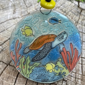 May include: A round glass ornament with a painted scene of a sea turtle swimming in blue water with coral, seaweed, and yellow fish. The ornament has a textured surface that resembles water droplets.