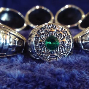 May include: A collection of silver rings with detailed engravings and a central green gemstone. The rings are displayed on a textured blue surface. The design includes intricate patterns and symbols, creating a striking visual effect.