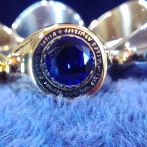 May include: A gold ring with a large blue gemstone set in the center. The ring has an inscription around the gemstone that reads "Lohia Jimmy O'Neil".