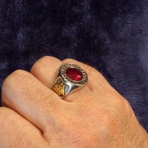 Könnte beinhalten: Ein silberner Ring mit einem roten Edelstein, der in einer goldfarbenen Fassung sitzt. Der Ring hat eine Band mit einem goldfarbenen Design und Runen um die Fassung.