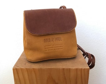 Op de afbeelding: Een klein bruin leren schoudertasje met een klep sluiting. De tas heeft een beige leren body en een bruine leren klep met de tekst "GOLD/PFEIL LUXURY IN LEATHER HANDMADE SINCE 1955 IN WEST GERMANY" erop gegraveerd.