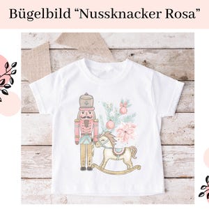 Könnte beinhalten: Weißes T-Shirt mit einer rosa und roten Illustration eines Nussknackers, der ein Schaukelpferd hält. Der Text "Bügelbild "Nussknacker Rosa"" befindet sich oben im Bild.