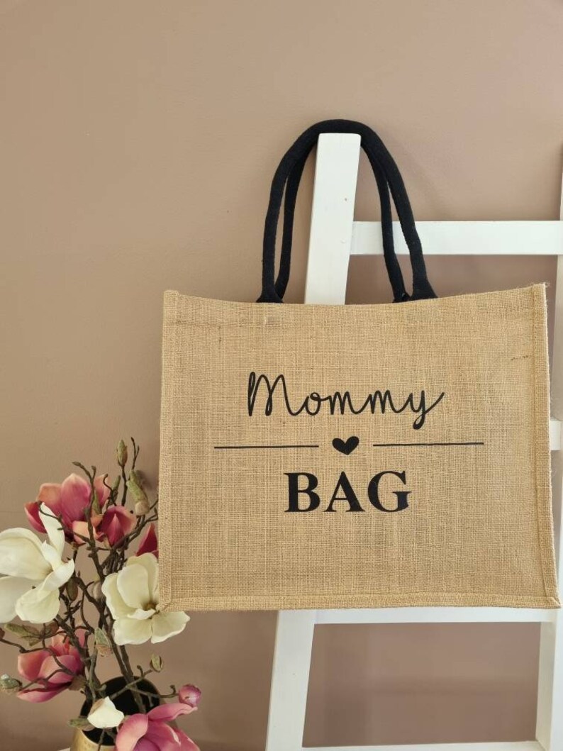May include: A brown burlap tote bag with black handles and the text "Mommy Bag" printed on the front in black.