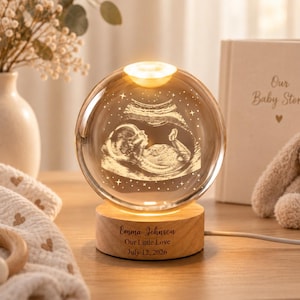 May include: A spherical glass lamp featuring an internal 3D laser-etched ultrasound image of a fetus. The glass sphere rests on a circular wooden base engraved with the text Emma Johnson, Our Little Love, July 12, 2026.