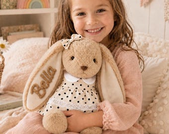 May include: A plush tan rabbit toy with long floppy ears, wearing a cream-colored dress with black polka dots, a white collar, and a lace trim. The name Bella is embroidered in brown thread on one ear, and a small matching polka dot bow is attached.