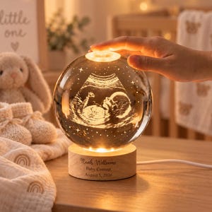 Könnte beinhalten: Ein kugelförmiges Glas-Nachtlicht mit einem 3D-lasergravierten Ultraschallbild eines Fötus im Inneren. Die Glaskugel steht auf einem runden Holzsockel, der mit dem Text Noah Williams, Baby Coming, August 5, 2026 graviert ist.