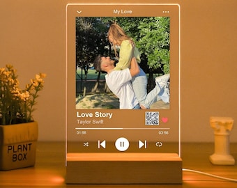 Könnte beinhalten: Ein Holz-Bilderrahmen mit einer klaren Acrylglasscheibe, auf der ein Foto eines Paares zu sehen ist. Der Rahmen verfügt über einen eingebauten Musikplayer mit Steuerelementen zum Abspielen, Pausieren und Überspringen. Das Display zeigt den Songtitel "Love Story" von Taylor Swift und einen Timer.