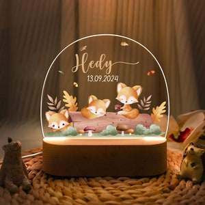 以下が含まれることがあります： 3匹のキツネと「Hedy」という名前と「13.09.2024」という日付が書かれた森のシーンが描かれた、透明なアクリル製のドームが付いた木製ナイトライトです。