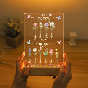 May include: A wooden light-up sign with a clear acrylic panel featuring a floral design and the text "First mommy - now nana". The sign is illuminated with warm white LED lights.