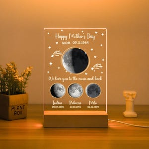 Peut inclure: Une veilleuse en acrylique personnalisée avec un motif de lune et d'étoiles. Le texte indique "Happy Mother's Day MOM 09.11.1964 We love you to the moon and back Indira 05.04.1990 Rebecca 22.10.1991 Milo 06.10.1999".