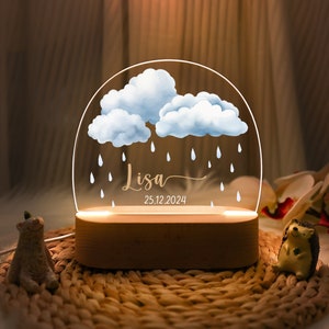 May include: A wooden night light with a clear acrylic insert featuring a blue watercolor cloud design with raindrops. The name "Lisa" and the date "25.12.2024" are inscribed on the base.