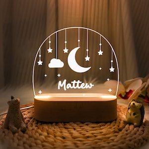 May include: A wooden night light with a white acrylic insert featuring a crescent moon, a cloud, and stars. The name "Matthew" is written below the moon and stars. The night light is illuminated with a warm white glow.
