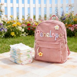 Può includere: Uno zaino rosa in velluto a coste con il nome "Penelope" ricamato in colori pastello. Lo zaino ha una piccola tasca frontale e un'applicazione a margherita. Una pila di pannolini è accanto allo zaino su una coperta a scacchi.