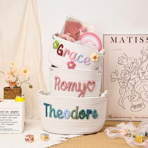 Puede incluir: Tres cestas de cuerda blanca apiladas, cada una con un nombre en hilo de colores: "Grace", "Romy" y "Theodore". La cesta superior contiene un arco&iacute;ris y otros art&iacute;culos. Acentos florales y una impresi&oacute;n de Matisse est&aacute;n en el fondo.