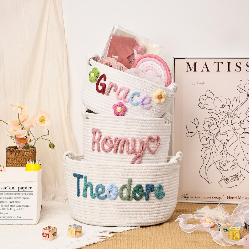 Puede incluir: Tres cestas de cuerda blanca apiladas, cada una con un nombre en hilo de colores: "Grace", "Romy" y "Theodore". La cesta superior contiene un arco&iacute;ris y otros art&iacute;culos. Acentos florales y una impresi&oacute;n de Matisse est&aacute;n en el fondo.
