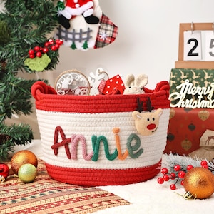 May include: A red and white rope basket with the name "Annie" in colourful letters. The basket is decorated with a reindeer and filled with Christmas ornaments. A Christmas stocking and gifts are in the background.