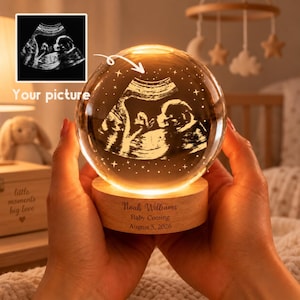 May include: A clear glass sphere lamp featuring an engraved ultrasound image inside, resting on a circular wooden base. The base is engraved with the text Noah Williams, Baby Coming, August 5, 2026, and emits a warm glow.