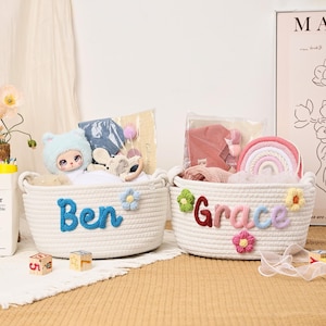 Puede incluir: Dos cestas de cuerda blancas, cada una con un nombre en hilo colorido: "Ben" y "Grace". Las cestas est&aacute;n llenas de juguetes y art&iacute;culos para beb&eacute;s, incluyendo una mu&ntilde;eca, un arco&iacute;ris y ropa. Flores decorativas adornan las cestas.