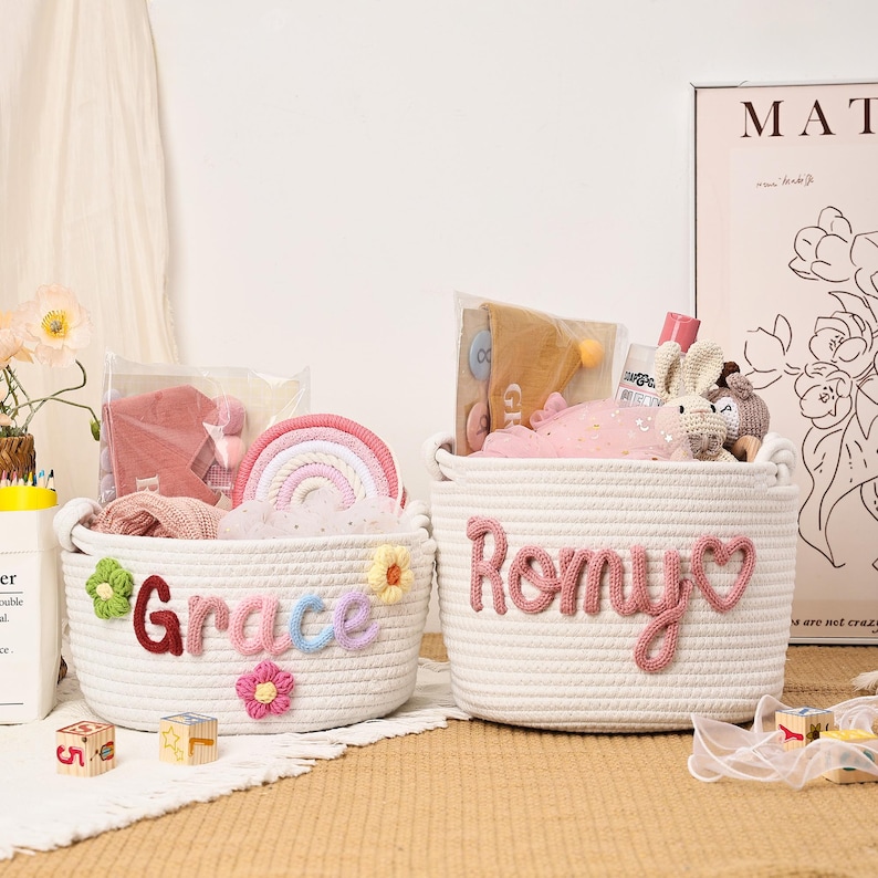 Puede incluir: Dos cestas de cuerda blanca, una con la etiqueta "Grace" con detalles florales y de letras coloridas, y la otra con la etiqueta "Romy" con letras rosas y un coraz&oacute;n. Ambas est&aacute;n llenas de juguetes y regalos, incluyendo un arco&iacute;ris, ropa y peluches.
