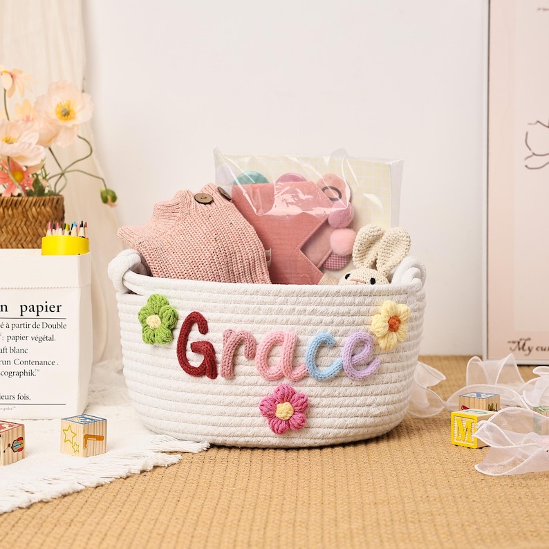 Puede incluir: Una cesta de cuerda blanca con el nombre "Grace" en letras coloridas y adornos florales. La cesta contiene un gorro de punto rosa, un coj&iacute;n en forma de estrella y un conejito de peluche. Un regalo ideal para un beb&eacute;.