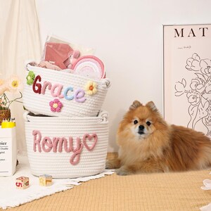 Puede incluir: Dos cestas de cuerda blanca, una con el nombre "Grace" y la otra con "Romy" en hilo de colores. La cesta "Grace" tiene adornos de flores. Un perro Pomerania, esponjoso y de color canela, est&aacute; acostado junto a las cestas.