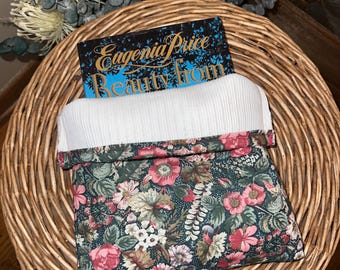 May include: A floral print fabric pouch with a white interior, holding a book titled "Eugenia Price Beauty from". The pouch features a dark green background with pink and white flowers. The items are on a woven basket.