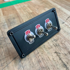 May include: A black panel featuring three toggle switches. Each switch has a sticker with "ON" in red and "OFF". The rectangular panel has rounded corners and is secured with four screws.