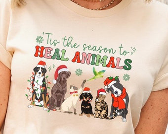 May include: Cream-colored t-shirt with the text "'Tis the season to HEAL ANIMALS" above a graphic of dogs and cats wearing holiday attire. The animals are arranged in a row, with festive decorations.