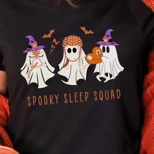 May include: Black t-shirt featuring three cartoon ghosts wearing witch hats and holding a pumpkin. The text "SPOOKY SLEEP SQUAD" is printed in orange. The shirt is a Halloween-themed design.