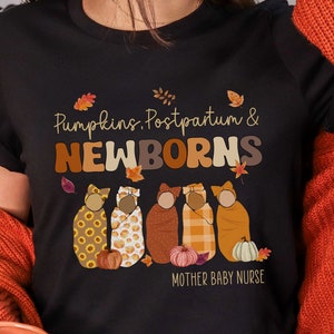 May include: Black t-shirt with the text "Pumpkins, Postpartum & NEWBORNS" in brown and orange lettering. The shirt features illustrations of swaddled babies, pumpkins, and autumn leaves. The words "MOTHER BABY NURSE" are printed below the illustrations.
