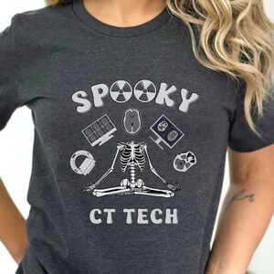 Peut inclure: T-shirt gris foncé avec l'inscription "SPOOKY CT TECH". Le motif présente un squelette en position de yoga entouré d'icônes d'imagerie médicale. Idéal pour Halloween ou un technicien en radiologie.