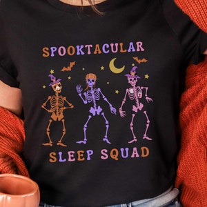 May include: Black t-shirt with the text "SPOOKTACULAR SLEEP SQUAD" and a graphic of three skeletons wearing witch hats. The skeletons are in orange, purple, and pink. Bats, stars, and a crescent moon are also featured in the design.