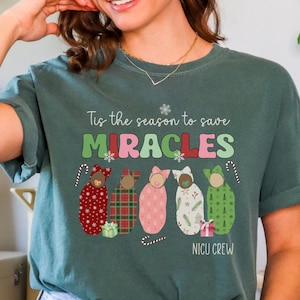May include: Dark green t-shirt with the text "Tis the season to save MIRACLES" and "NICU CREW". The shirt features illustrations of swaddled babies in festive patterns and candy canes, perfect for the holiday season.