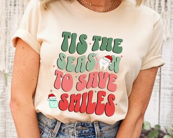 May include: Cream-colored t-shirt with the text "Tis the Season to Save Smiles" in green, pink, and red. The design includes a tooth wearing a Santa hat and a cartoon tooth with a Santa hat. The shirt is paired with blue jeans.