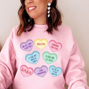 May include: A pale pink sweatshirt with heart-shaped candies in various colours. The candies have phrases such as "Breathe" and "Lunging for You". The sweatshirt is a long-sleeved crew neck style.