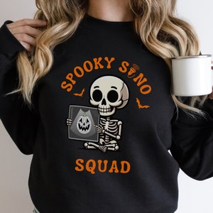 Peut inclure: Sweat-shirt noir avec l'inscription "SPOOKY SONO SQUAD" en orange. Le motif représente un squelette de dessin animé tenant une image d'échographie d'une citrouille. Des chauves-souris sont également incluses dans le motif.