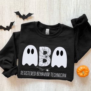 May include: Black sweatshirt with a Halloween theme. The design features two white ghost figures flanking the letters "ABA" with a spider web design in the middle. The text "REGISTERED BEHAVIOR TECHNICIAN" is below the letters. Decorative bats and a small pumpkin are also present.