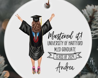 May include: White round ornament with a black and white illustration of a graduate wearing a cap and gown. The text "Mastered it! University of Hartford M.Ed Graduate Class of 2024 Andrea" is printed on the ornament.