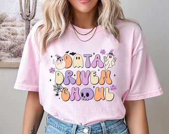 May include: Light pink t-shirt with the text "DATA DRIVEN GHOUL" in a retro font, featuring ghost and skull graphics. The shirt has a relaxed fit and short sleeves, perfect for Halloween or spooky season.